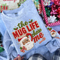 A light heather blue crewneck sweatshirt featuring a large, festive graphic print of a winking Santa Claus face surrounded by the text 'the MUG LIFE chose me' in red and green lettering with holly and candy cane accents. The sweatshirt is styled in a cozy holiday flat-lay setting with gift boxes and ribbons.