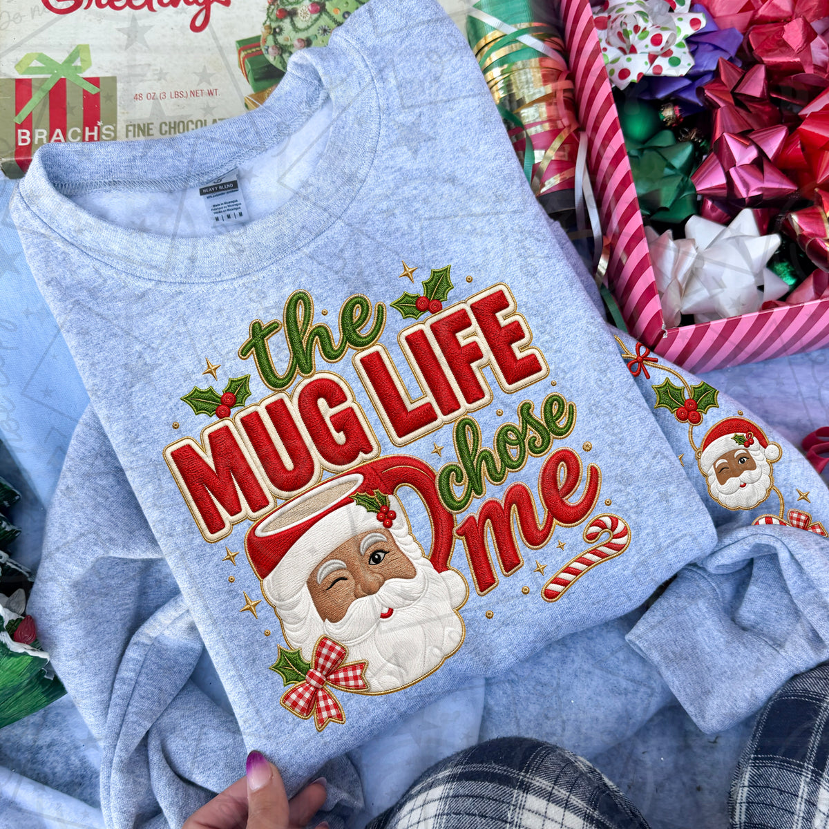 A light heather blue crewneck sweatshirt featuring a large, festive graphic print of a winking Santa Claus face surrounded by the text 'the MUG LIFE chose me' in red and green lettering with holly and candy cane accents. The sweatshirt is styled in a cozy holiday flat-lay setting with gift boxes and ribbons.